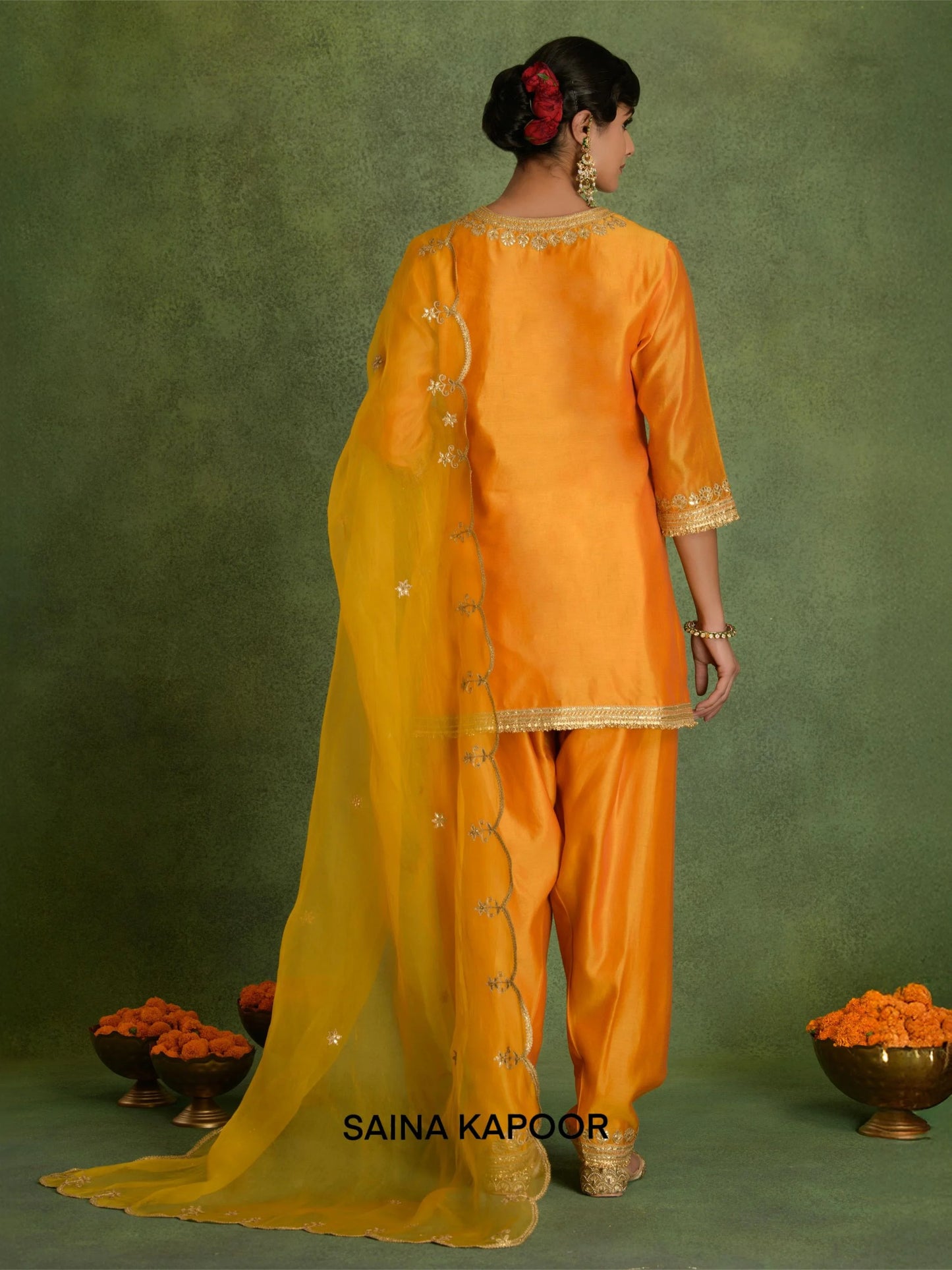Back view of orange marigold Chanderi short A-line kurta set for women showing graceful A-line hem flare, three-quarter gota embroidered sleeve cuffs, contrasting mustard scalloped organza dupatta & matching dhoti salwar with embroidered ankle cuffs — Saina Kapoor