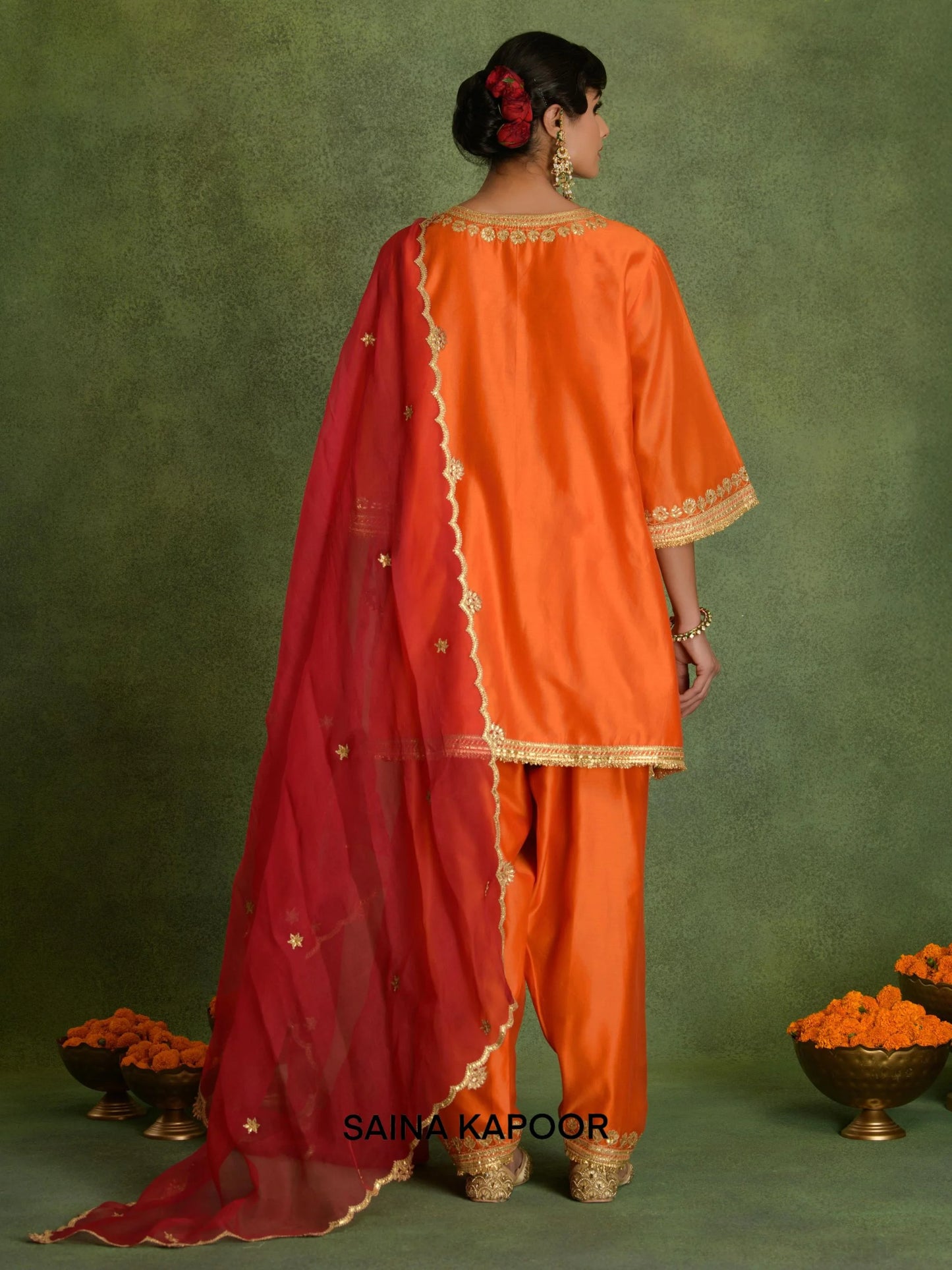 Back view of deep orange Chanderi short kurta set for women showing three-quarter gota embroidered sleeve cuffs, contrasting red scalloped organza dupatta & matching dhoti salwar with embroidered ankle cuffs — Saina Kapoor
