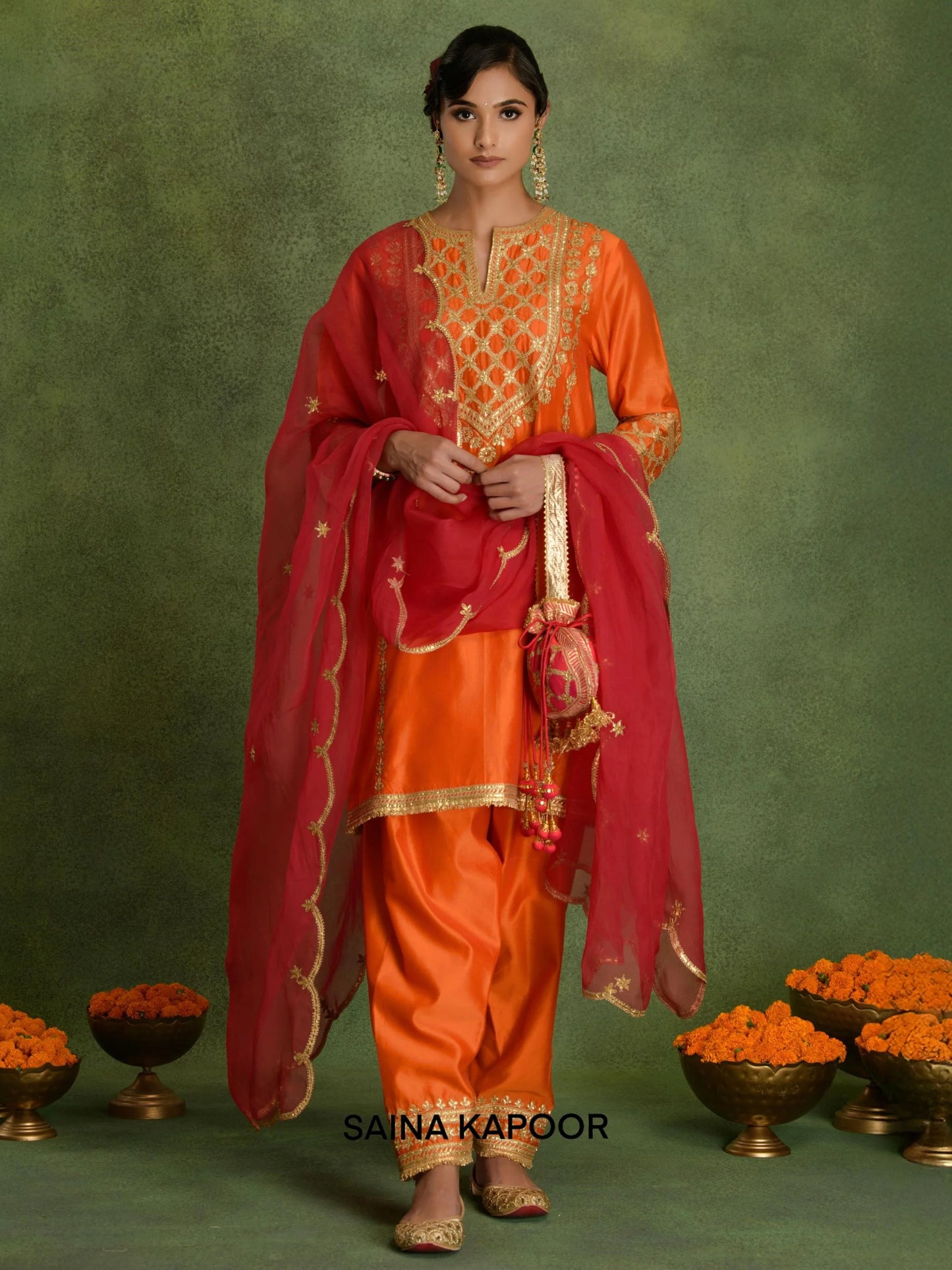 Deep orange Chanderi short kurta DEBANJALI with gold gota patti geometric trellis embroidered V-notch yoke, matching dhoti salwar with embroidered ankle cuffs, contrasting red dupatta & pink potli bag — Saina Kapoor