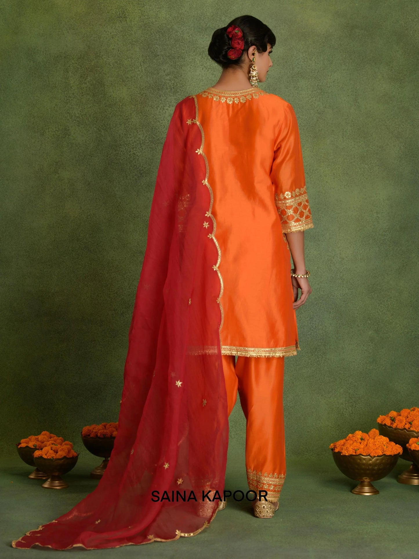 Back view of deep orange Chanderi short kurta set for women showing three-quarter gota embroidered sleeve cuffs, contrasting red scalloped organza dupatta & matching dhoti salwar with embroidered ankle cuffs — Saina Kapoor
