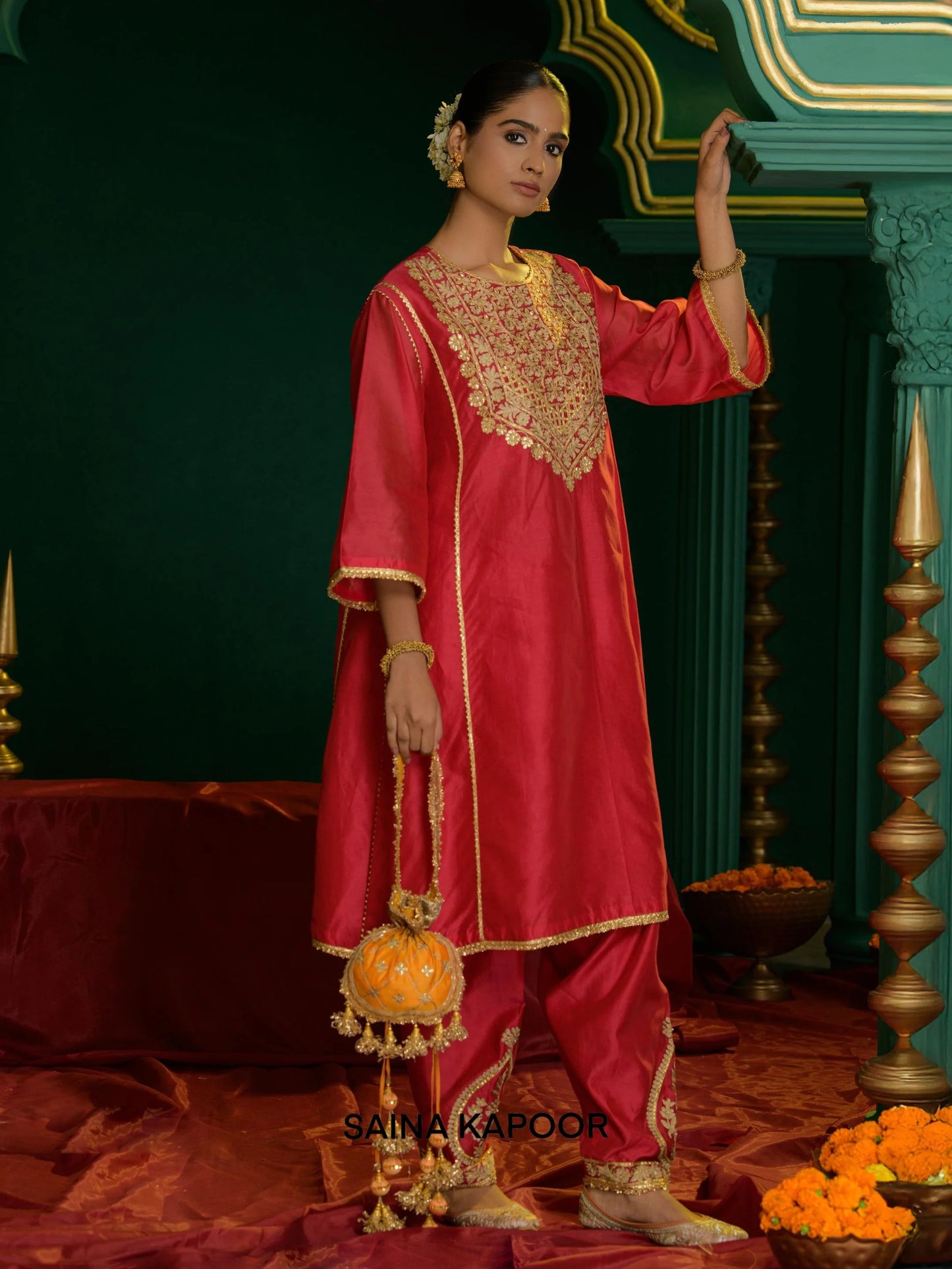 Side angle of coral red Chanderi long choga ANURA showing open-front silhouette, gota patti embroidered yoke, dhoti salwar with embroidered ankle cuffs & orange potli bag — Saina Kapoor